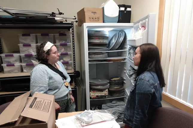 Two people standing talking in collections storage room next to open freezer with film canisters inside